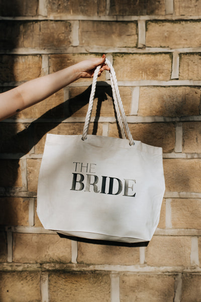 A white tote bag with 'THE BRIDE' text on it, held against a brick wall background.